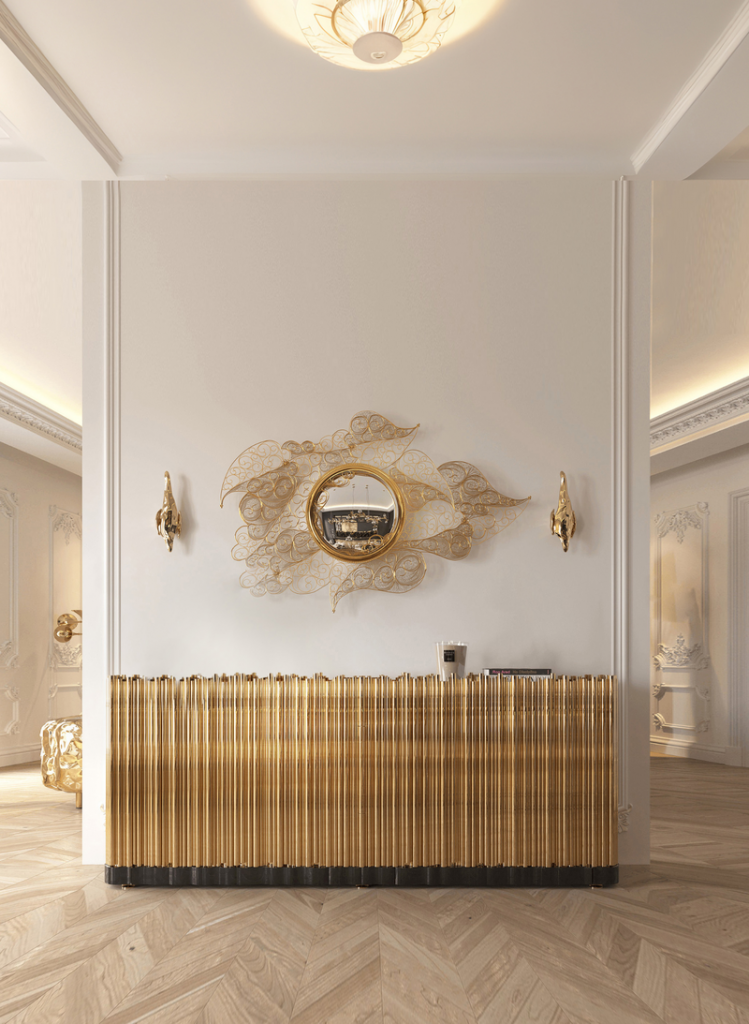 Gold filigree-framed mirror shaped like flowing leaves, mounted above a ribbed gold console table with matching wall sconces in a bright herringbone-floored hallway, luxury design.
