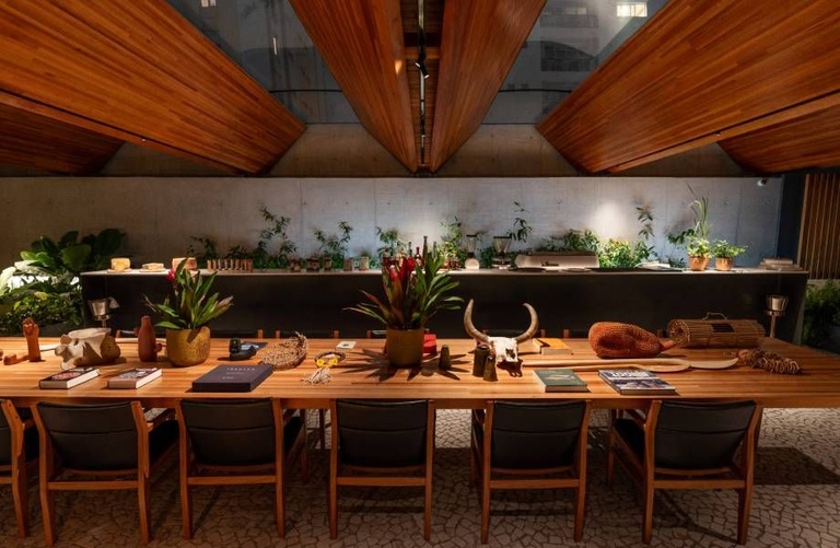 Mesa comunitária de madeira com cadeiras de couro sob teto triangular de madeira, decorada com plantas, livros e objetos artesanais - melhores restaurantes em São Paulo