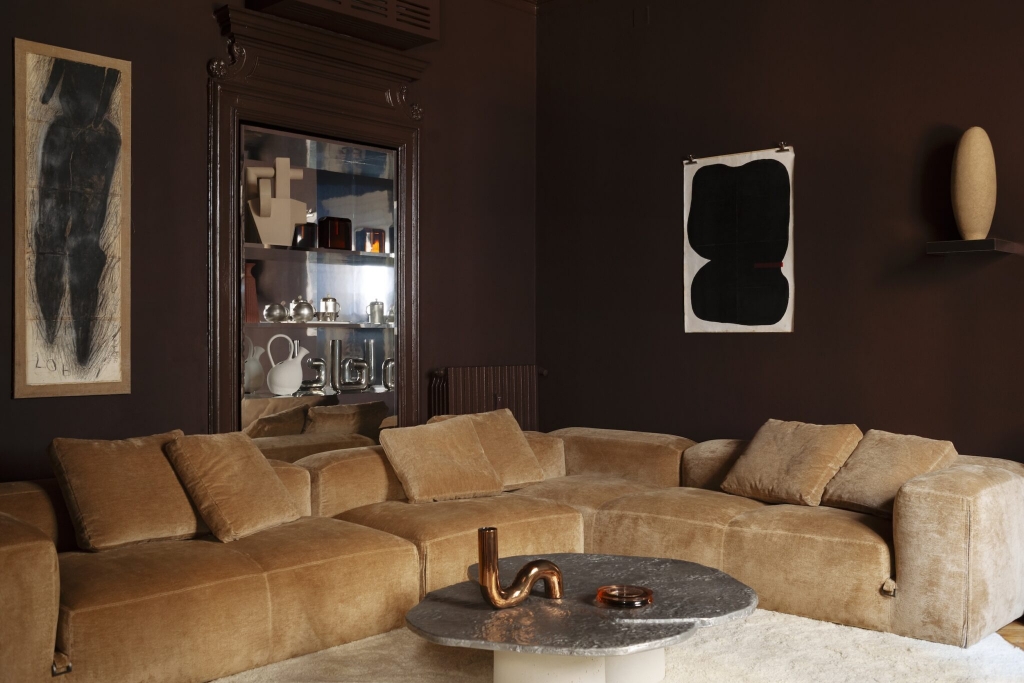Moody dark brown living room with a camel chenille sectional sofa, sculptural bronze and glass coffee table, framed abstract artwork, and a mirrored display cabinet showcasing ceramics. interior designers