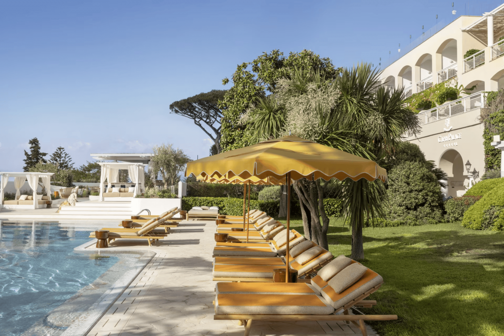 Luxury pool deck with yellow umbrellas, wooden lounge chairs, and lush palm trees. Old Money Hotels in Italy