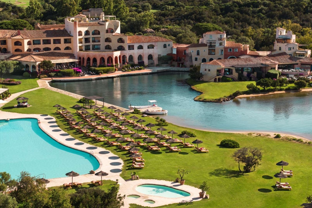Aerial view of a Sardinian resort complex with pools, sun loungers, and a lagoon surrounded by terracotta-roofed buildings