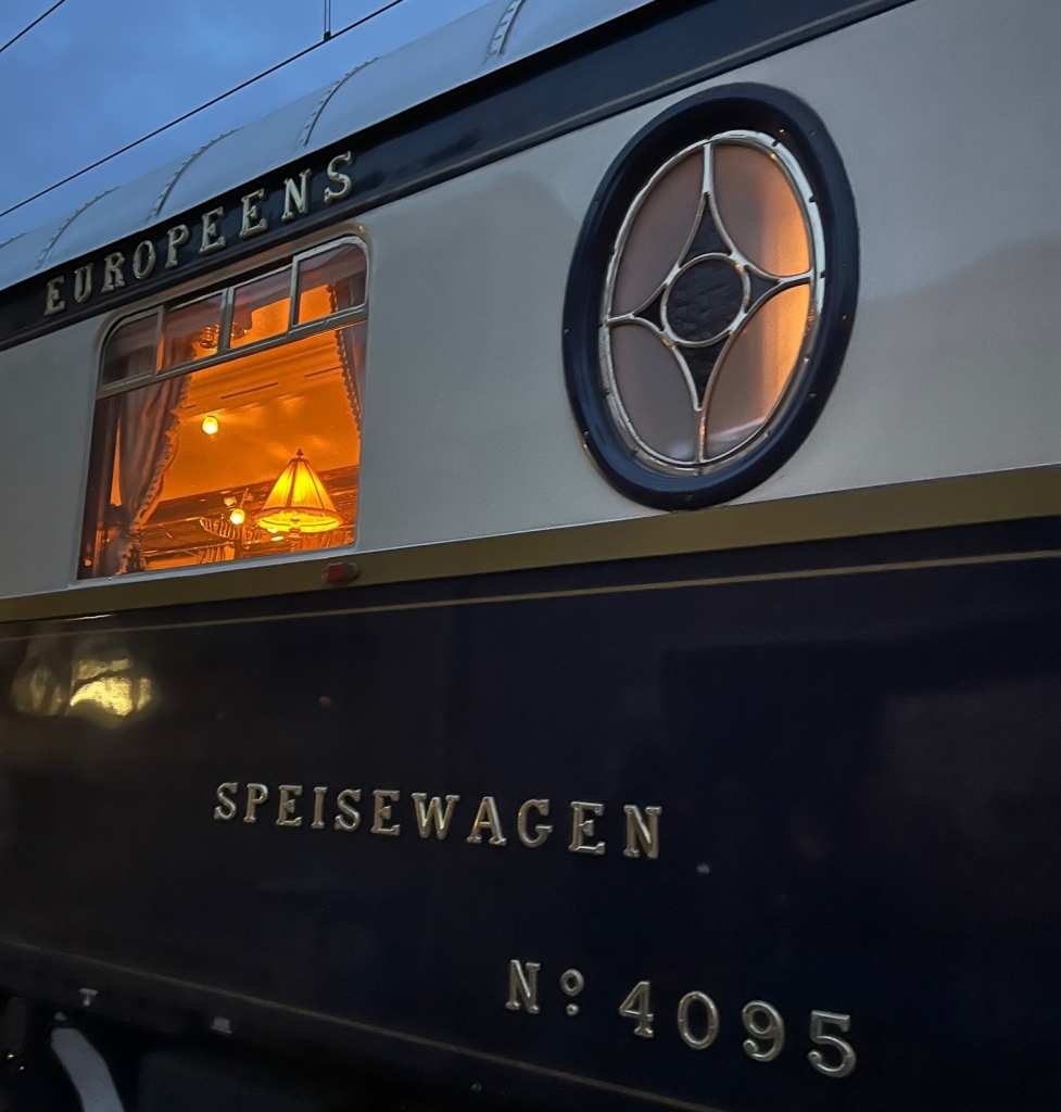 Evening exterior shot of a dining car labeled "Européens Speisewagen No. 4095" with a warmly lit window and stained-glass oval -Venice simplon-orient-express