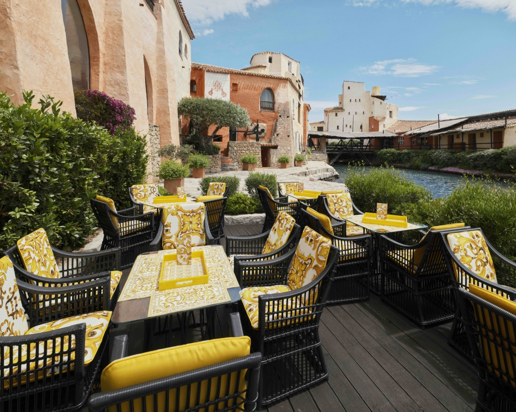 Outdoor dining terrace with wicker chairs upholstered in yellow majolica-print fabric, overlooking a canal and terracotta buildings