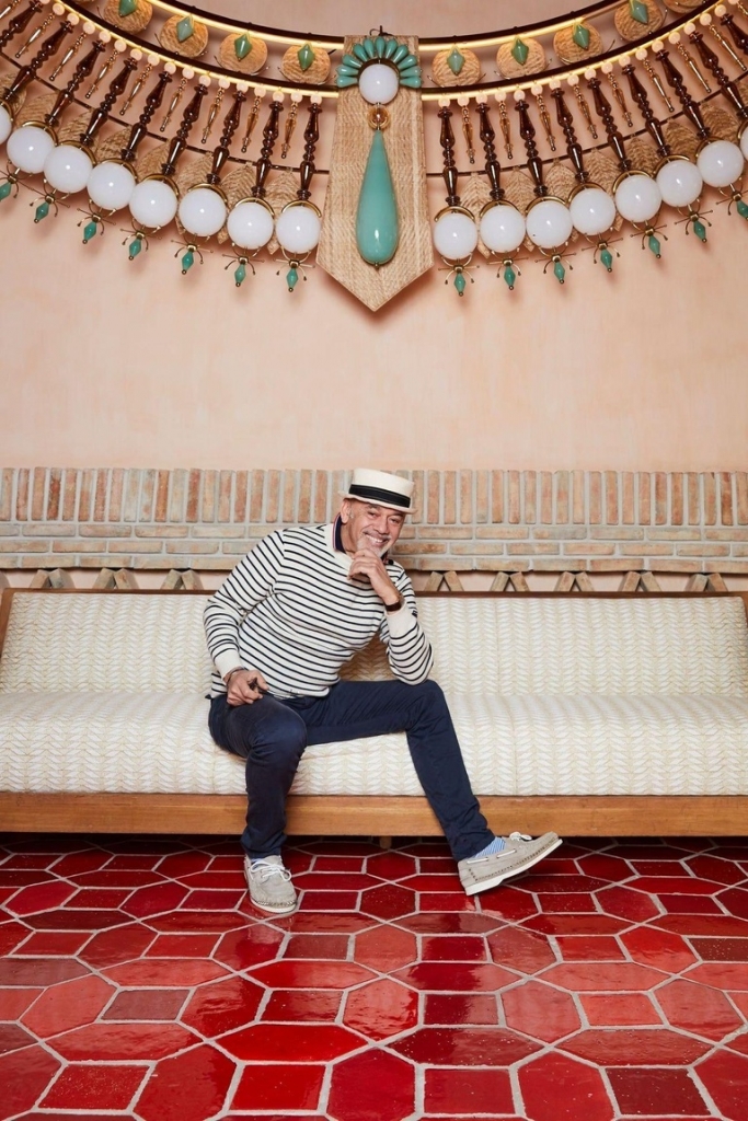 Portrait of a man in a striped sweater and boater hat sitting on a bench beneath an ornate beaded chandelier - christian Louboutin villas