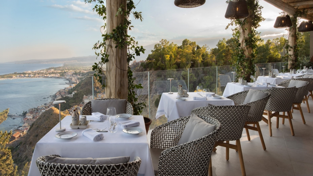 Hillside terrace dining area with patterned chairs and white-clothed tables overlooking the coastline and sea at dusk -four seasons hotel taormina