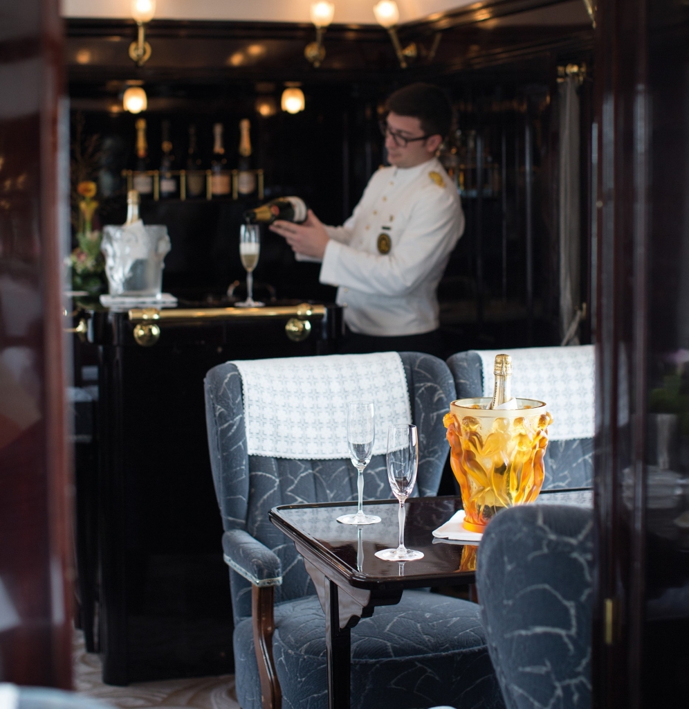 Steward in white uniform pouring champagne at a bar cart in a train lounge, with two flutes and an ice bucket on a table in the foreground -Venice simplon-orient-express
