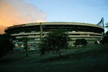 Football stadium seen at dusk, with smoke in the background and trees in the foreground -top places to live in São Paulo