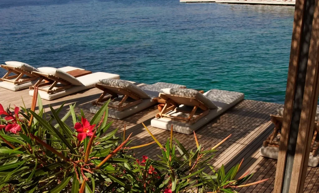 Wooden sun loungers on a deck by crystal-clear water, with flowers in the foreground -luxury beach clubs in Europe