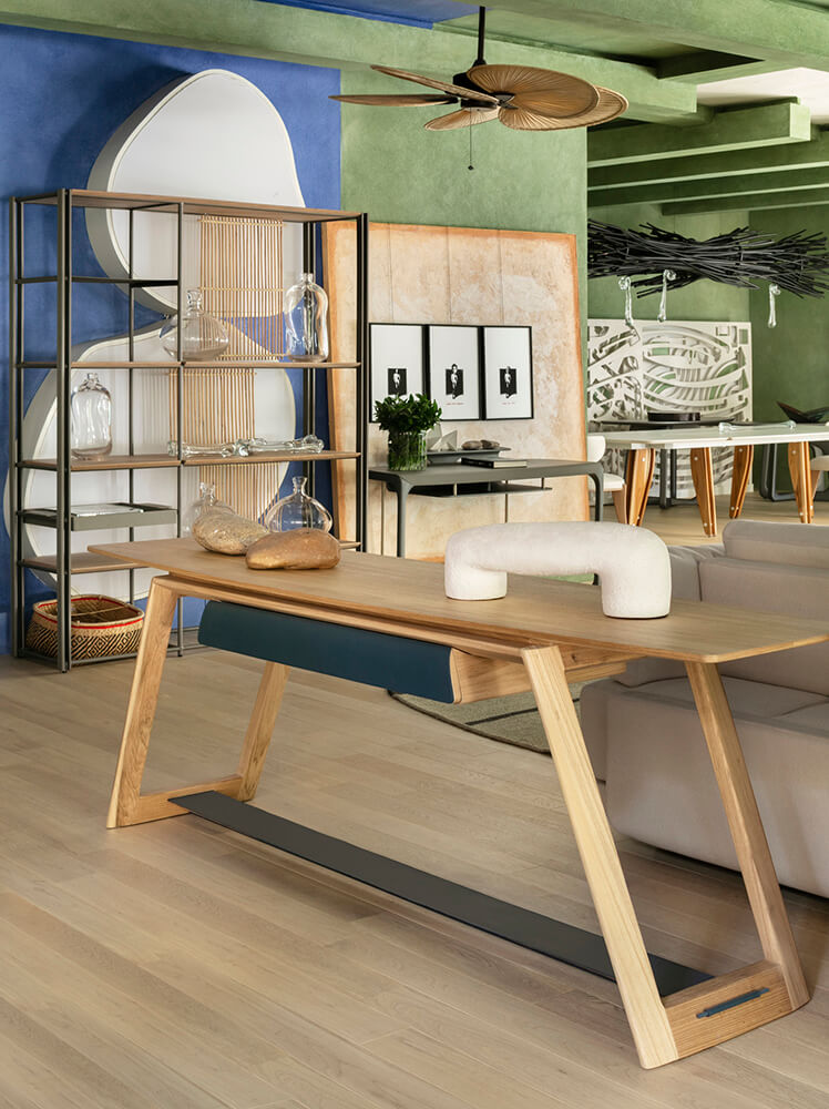 Wooden console table with a blue drawer accent in front of a shelving unit and mirror art piece -top showrooms in São Paulo