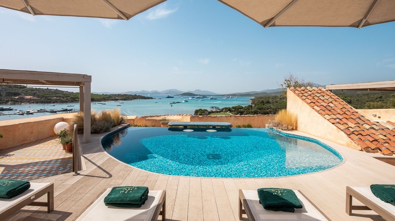 Infinity pool with a swim-up bar overlooking a scenic bay, flanked by lounge chairs with green towels