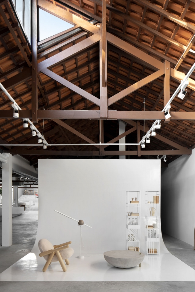 Interior showroom space with exposed wooden roof trusses and terracotta tiles above a white photography backdrop with furniture displays -showrooms in Brazil