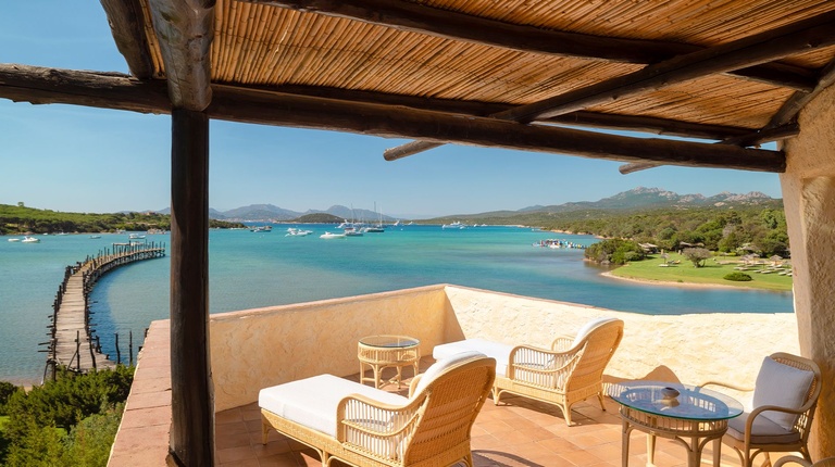Private balcony with rattan lounge chairs under a thatched pergola, overlooking a turquoise bay dotted with boats