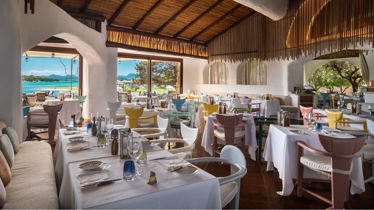 Bright restaurant dining room with pastel-colored chairs, white tablecloths, and arched windows overlooking the sea