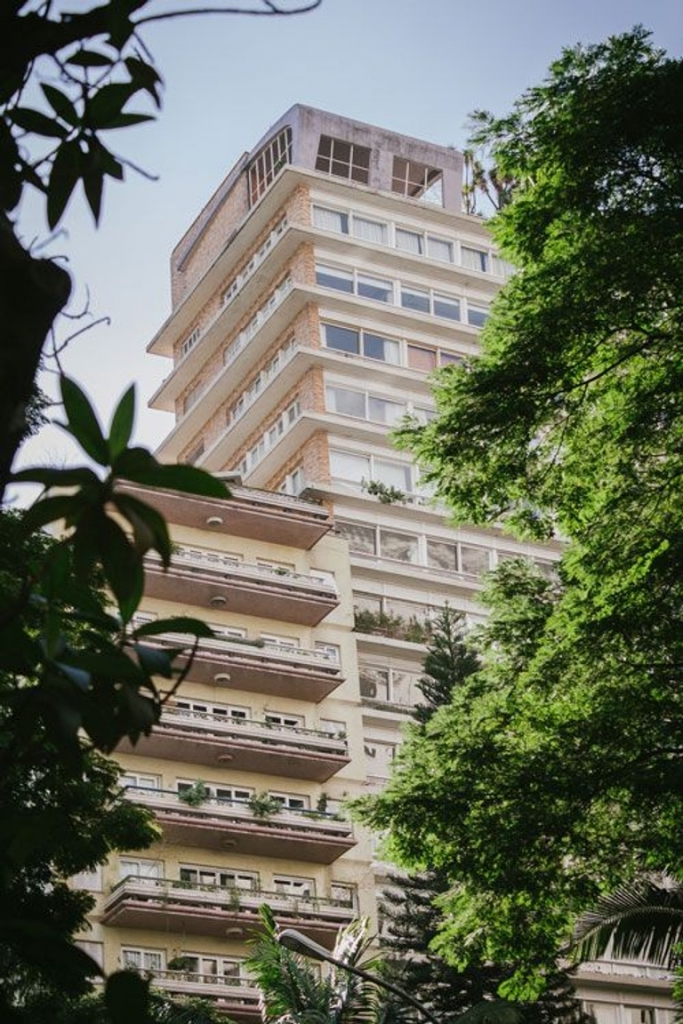 Low-angle view of a tall residential building with balconies, framed by green trees -top places to live in São Paulo
