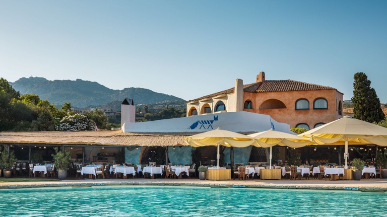 Poolside view of a Mediterranean beach club restaurant with terracotta buildings, yellow umbrellas, and mountains in the background