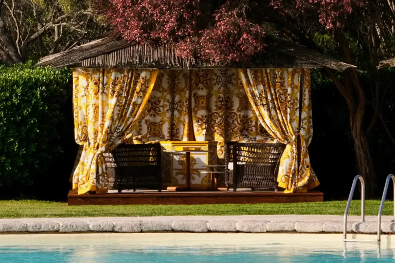 Poolside cabana draped with yellow and gold floral curtains, shaded by blossoming trees