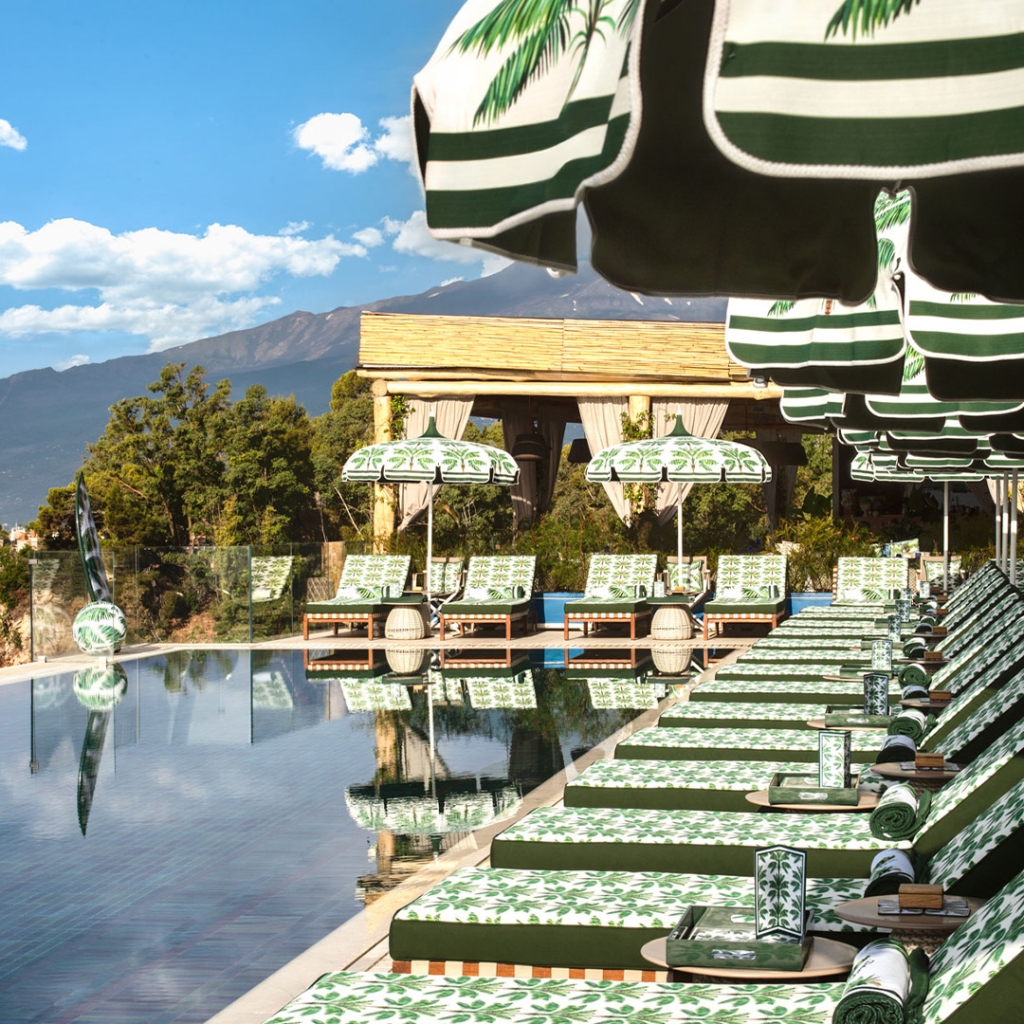 Row of green palm-print umbrellas and matching sun loungers beside an infinity pool with a palm tree and coastal town in the background -four seasons hotel taormina