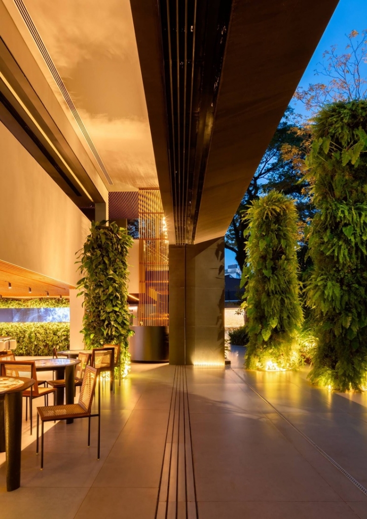 Outdoor showroom corridor with illuminated vertical garden columns and dining tables at twilight -showrooms in Brazil