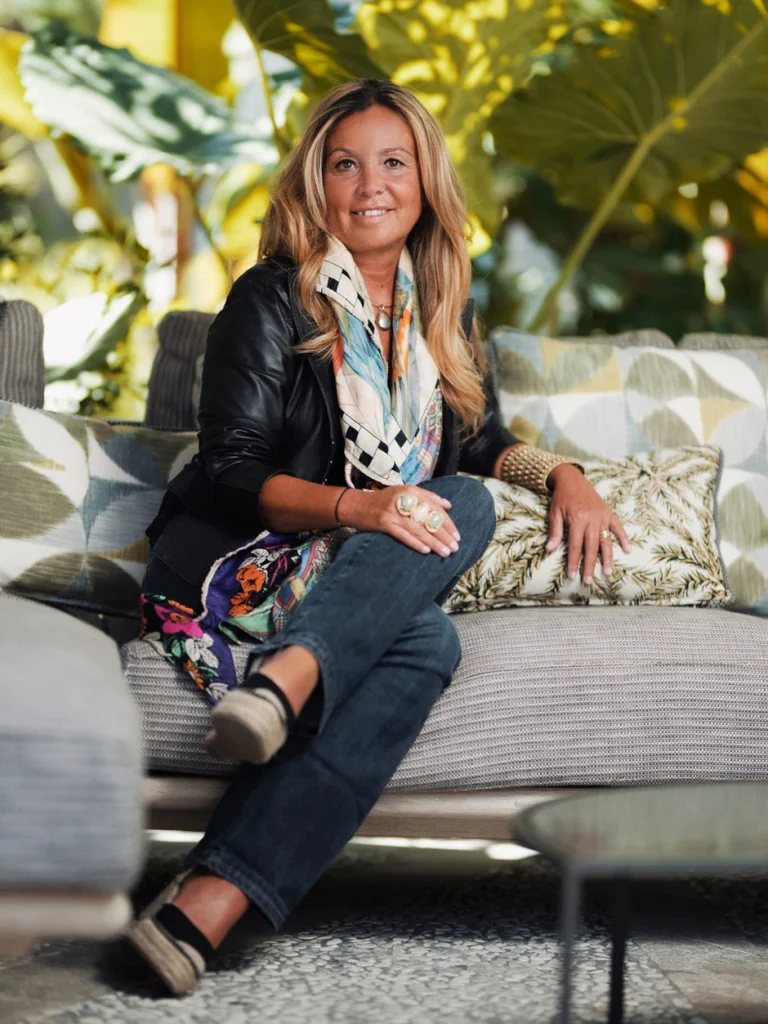 Portrait of a smiling woman in a leather jacket and patterned scarf seated on an outdoor sofa surrounded by greenery -four seasons hotel taormina