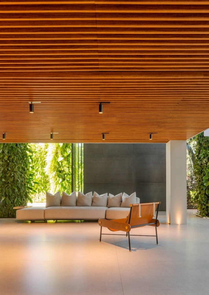 Spacious lounge area with a slatted wood ceiling, a beige sofa, and a camel leather chair beside glass walls with vertical gardens -showrooms in Brazil