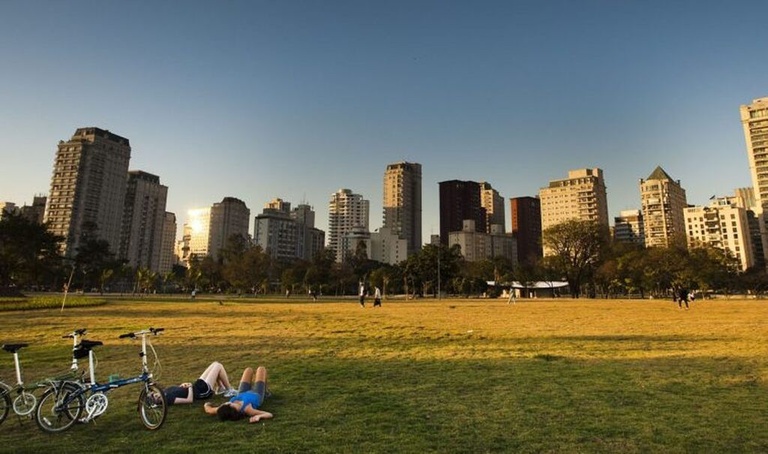 Two people lying on the grass in an urban park at sunset, with bicycles and a skyline of skyscrapers in the background -top places to live in São Paulo