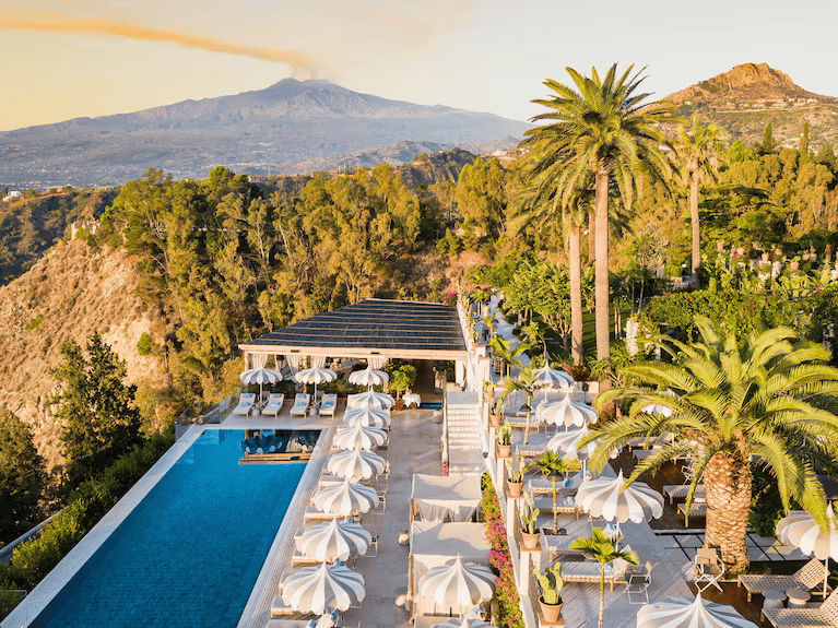 Resort swimming pool and terrace overlooking green hills with Mount Etna in the background. Old Money Hotels in Italy