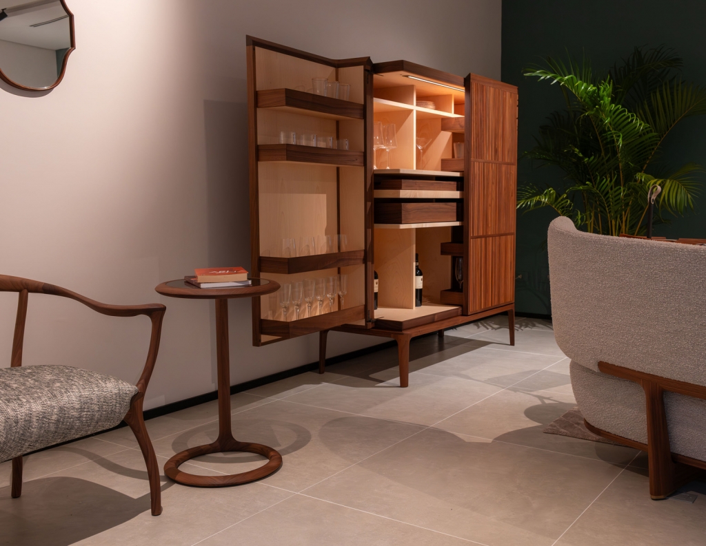Open wooden bar cabinet displaying glassware, wine bottles, and drawers beside an armchair and side table -top showrooms in São Paulo