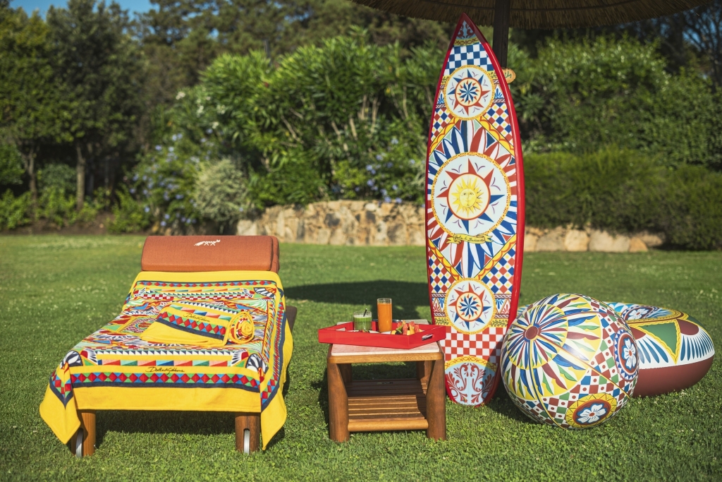 Poolside setup with a painted surfboard, decorative beach ball, and lounger featuring Dolce & Gabbana Carretto-print textiles