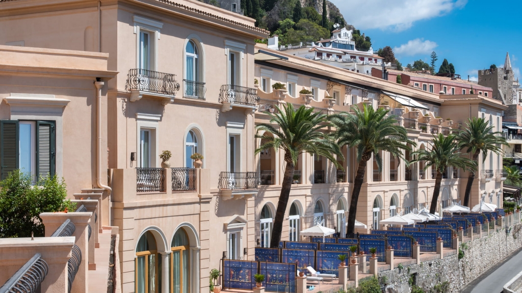 Elegant peach-toned hotel facade with wrought-iron balconies, palm trees, and blue tiled terrace panels along a hillside road -four seasons hotel taormina