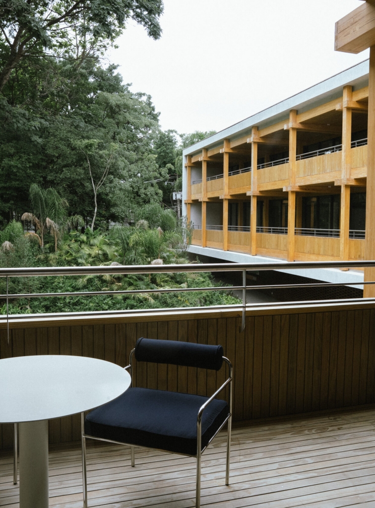 Wooden balcony with a black lounge chair and round table, overlooking a building with timber-clad balconies and lush greenery -showrooms in Brazil