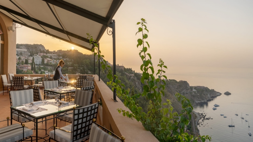 Terrace restaurant with striped chairs being set for dinner at sunset, overlooking a coastal cliffside and marina -four seasons hotel taormina