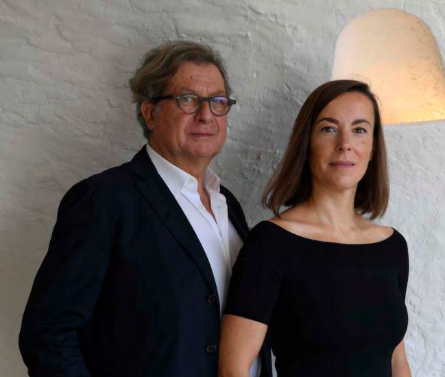 Portrait of a man and a woman standing together against a whitewashed wall, both dressed in dark clothing