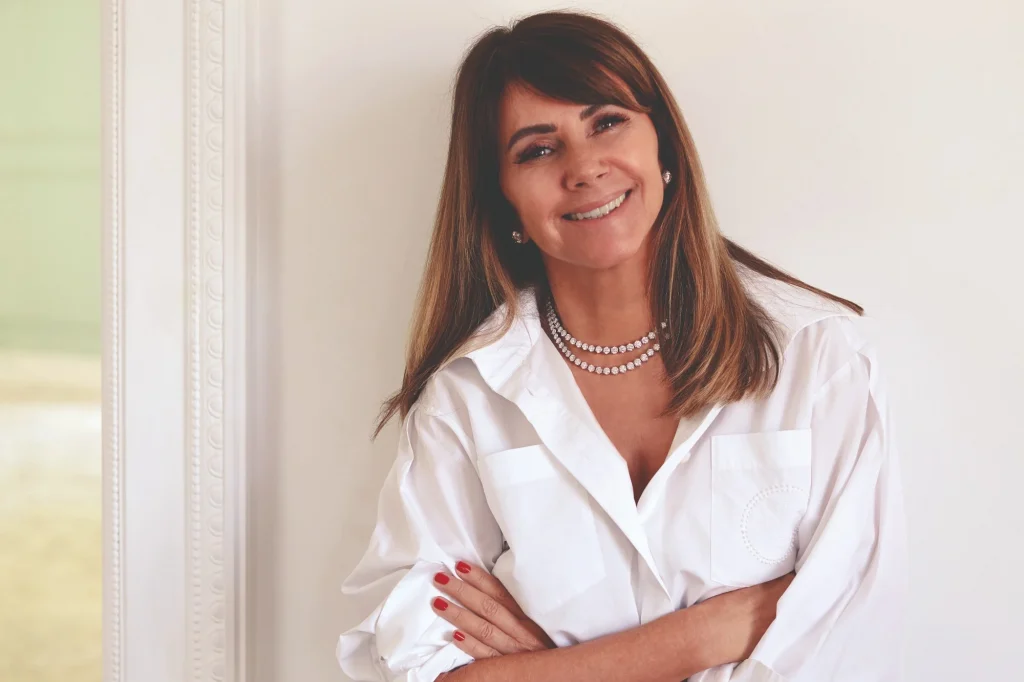 Tania Bulhões sorrindo com blusa branca social e colar de brilhantes.