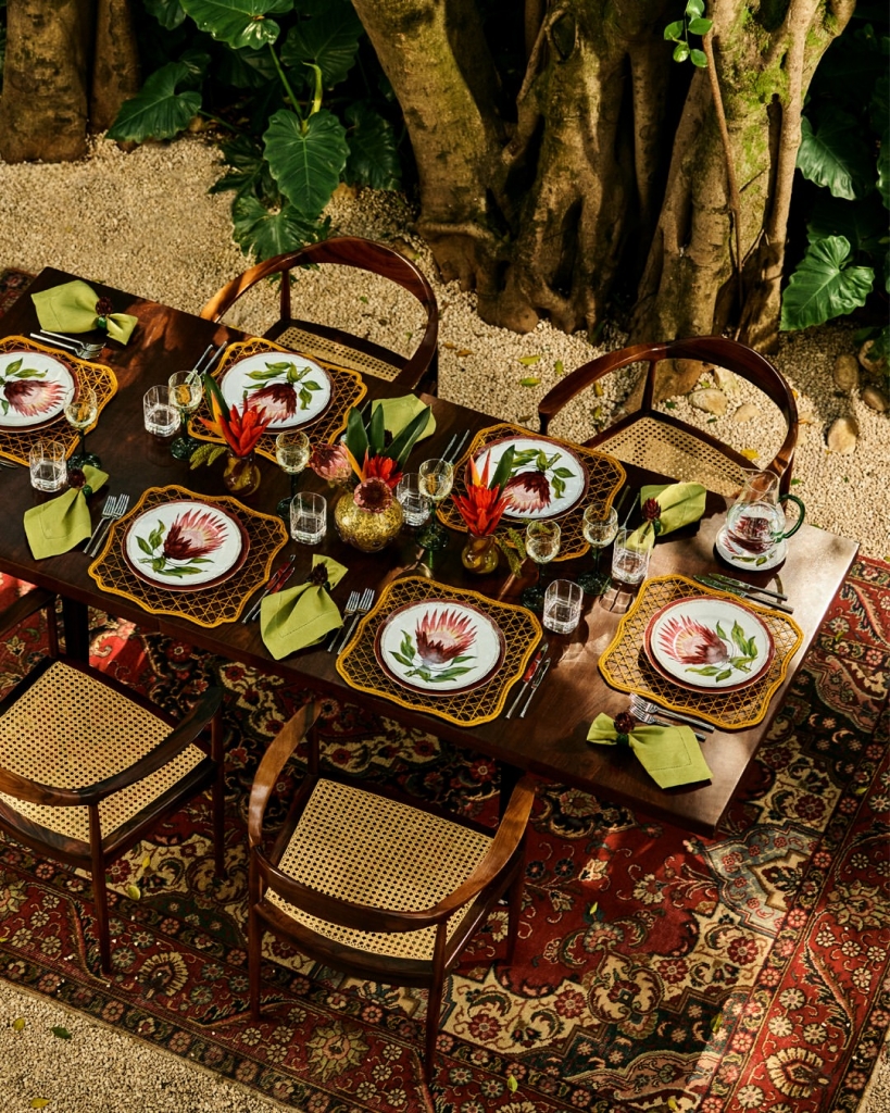 Mesa de jantar de madeira ao ar livre com jogos americanos amarelos e pratos da Coleção Botânica.