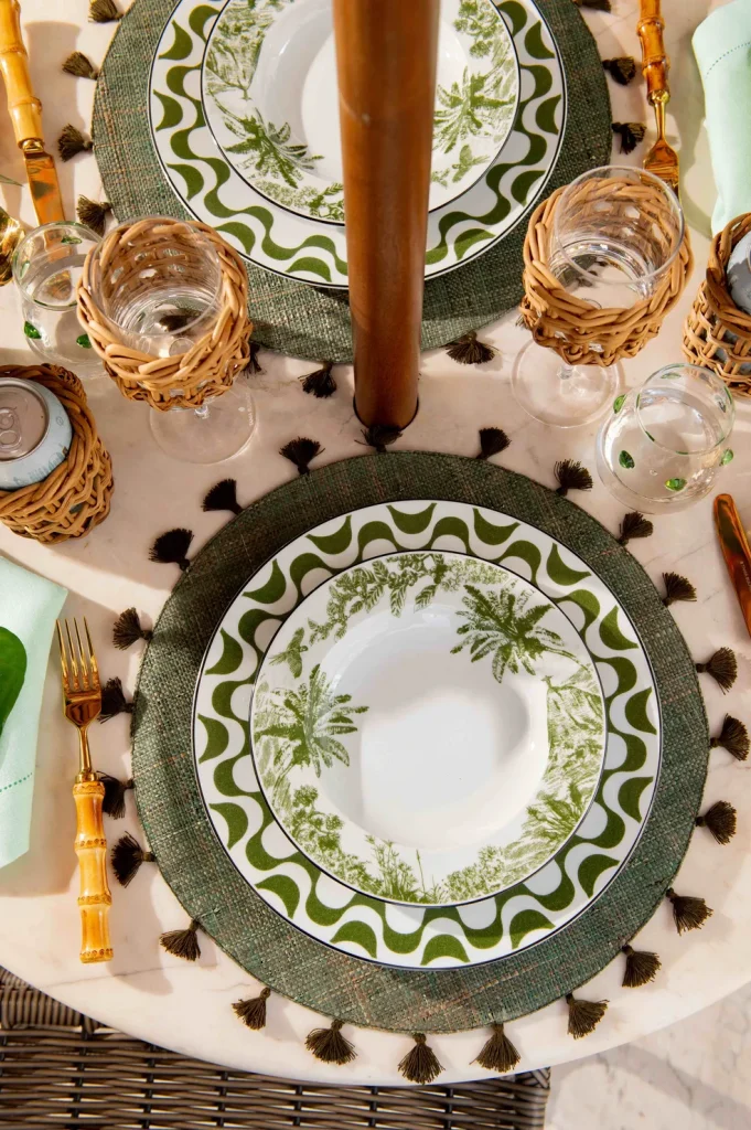 Mesa posta com placemat verde, pratos decorados com estampa tropical de palmeiras e talheres com cabo de bambu.