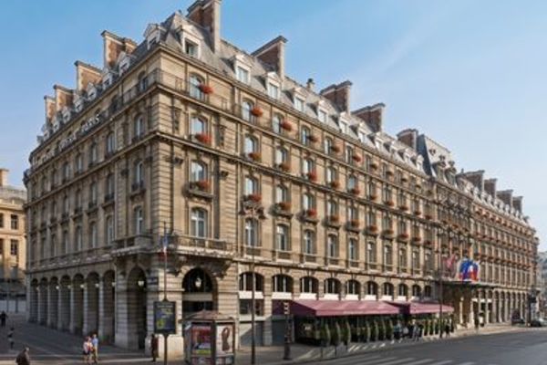 An exterior view of a historic luxury hotel building in Paris featuring classic Haussmann architecture, stone facades, rows of uniform windows with window boxes, arched ground-floor colonnades, and outdoor awnings. Paris design week.