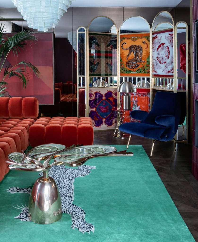 Eclectic living room with rust-orange tufted sofa, navy velvet armchair, emerald rug and mirrored folding screen with animal-print scarves - fall winter 2027 trends