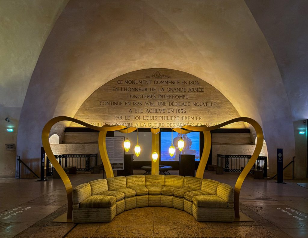 A curved beige upholstered sofa placed beneath a large golden sculptural arch inside a vaulted historic stone room at paris design week