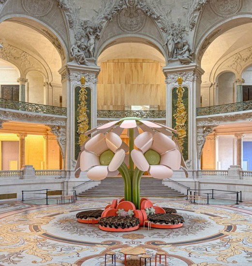A monumental pink flower sculpture installation with green stems and cushioned seating at its base, displayed in the grand hall of an ornate historic building with classical architecture, arches, and intricate stone carvings at paris design week