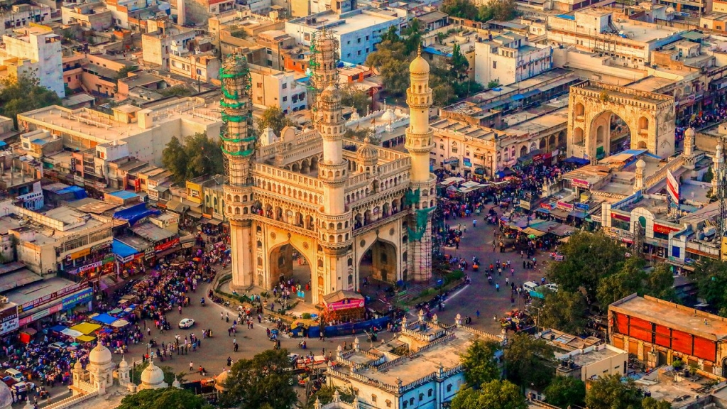 An aerial high-angle view of the historic Charminar monument in Hyderabad, surrounded by bustling streets filled with crowds of people, market stalls, and surrounding urban architecture. design democracy