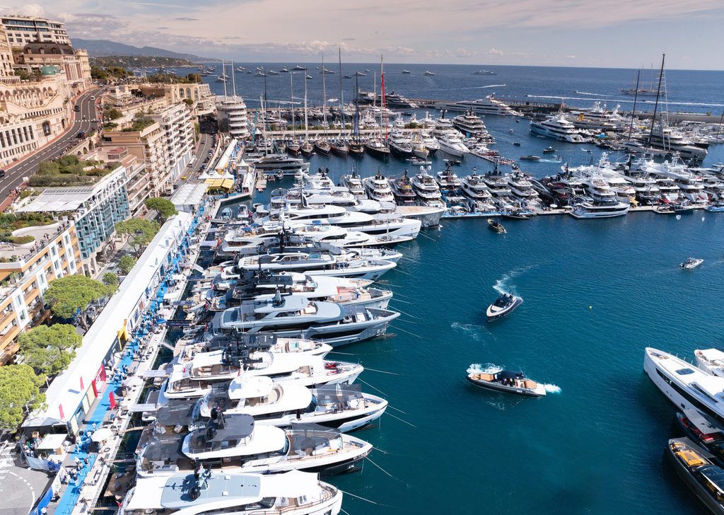 High-angle drone shot of a busy harbor showcasing numerous moored luxury yachts and tenders cruising on blue water next to coastal city buildings, Monaco Yacht Show 2026