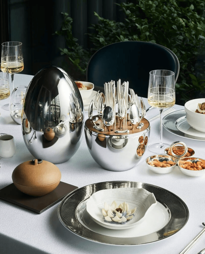 A formal dining table set with white tablecloths, fine silver tableware, a large silver egg-shaped container holding cutlery, wine glasses, and various bowls of snacks. Paris design week.