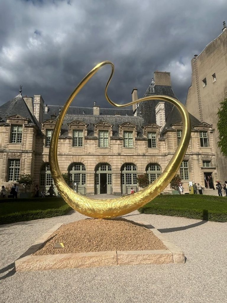A large, abstract golden loop sculpture installed on a gravel base in the courtyard of a historic classical French stone building under a cloudy sky at paris design week