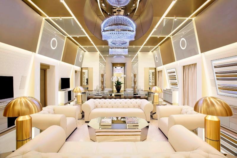 A symmetrical, ultra-luxurious living room featuring gleaming cream-colored leather sofas, gold-accented geometric coffee tables, large golden mushroom-shaped table lamps, and a cascading multi-tiered crystal chandelier, all grouped under most prestigious hotel suites