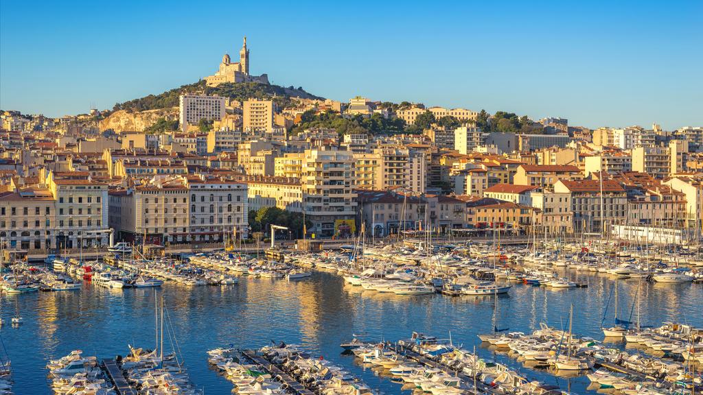 A panoramic cityscape of Marseille, France, taken from an elevated position, capturing the Old Port filled with hundreds of boats. The iconic Notre-Dame de la Garde basilica is perched prominently on the hill overlooking the densely packed buildings with terracotta roofs. The water of the port is calm, reflecting the golden light of the sun. Cannes Yachting Festival.