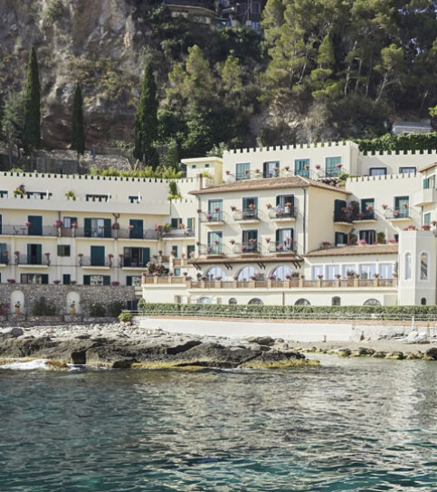 A Mediterranean-style resort built into a steep hillside, with cream-colored, multi-story buildings and terracotta roofs perched directly above rocky outcrops and the clear blue sea. Luxury hotel chains.