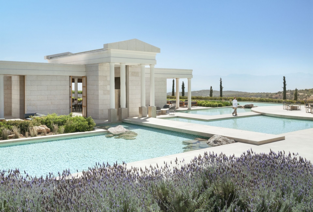 A modern building with classical architectural elements like columns and a pediment, situated beside a series of reflection pools, with fields of purple lavender in the foreground and distant hills under a clear sky. Luxury hotel chains.