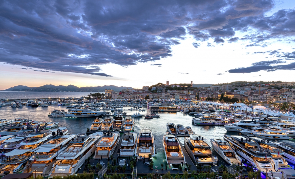 An aerial view of the bustling Vieux Port de Cannes at twilight, packed with hundreds of luxury superyachts. The city of Cannes and the surrounding hills are visible in the background under a cloudy sky, with lights beginning to glow on the boats and docks. Cannes Yachting Festival.
