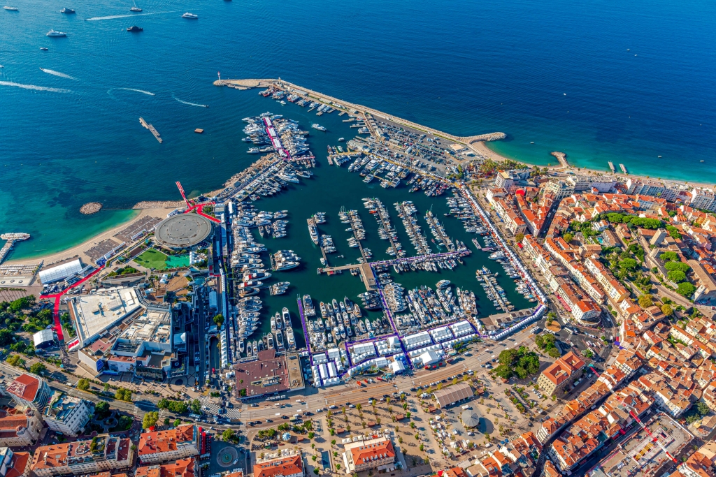 A high-angle aerial photograph of the entire Vieux Port de Cannes and surrounding city during the day. The marina is completely filled with a dense grid of yachts and superyachts. White event tents are set up along the quays. The red-tiled roofs of Cannes extend into the background, with the Palais des Festivals and beaches visible on the left, and a long breakwater on the right. Cannes Yachting Festival.