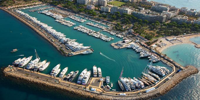 An aerial perspective of a large marina, possibly Port Pierre Canto, viewed from the east. The breakwater encloses a basin packed with various sized yachts and sailing boats, with a smaller outer harbor. Waterfront apartment buildings and a sandy beach line the shore. The water is a deep blue. Cannes Yachting Festival.